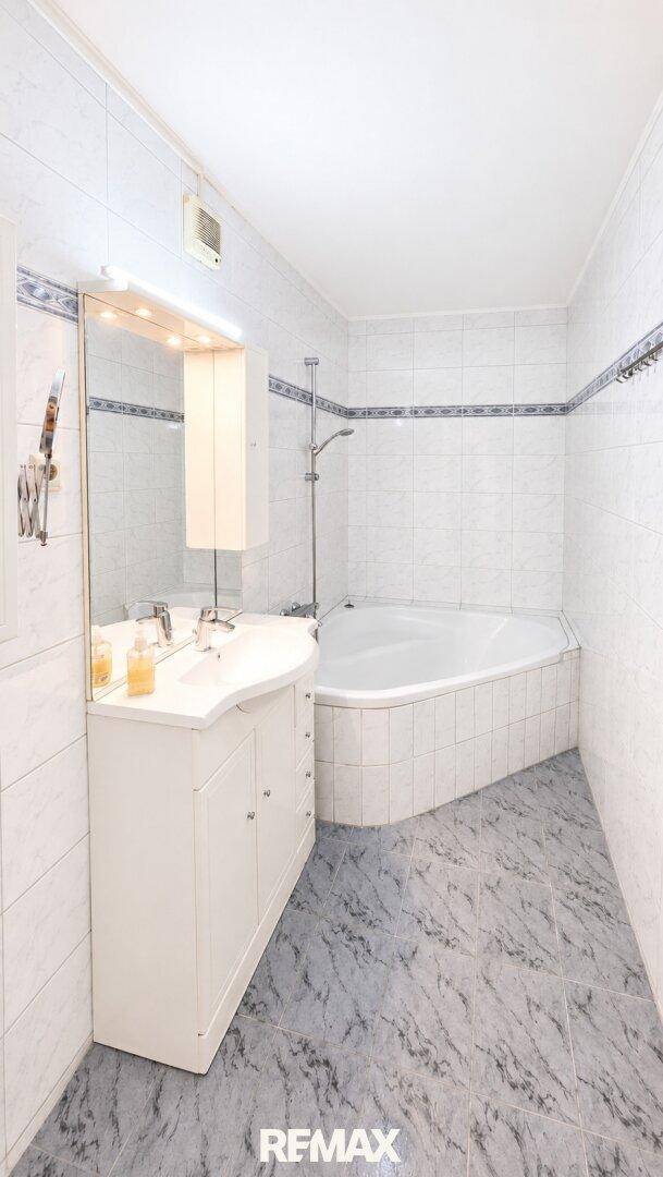 Badezimmer mit Eckbadewanne, Einzelwaschbecken und Spiegelschrank, hell gefliest.