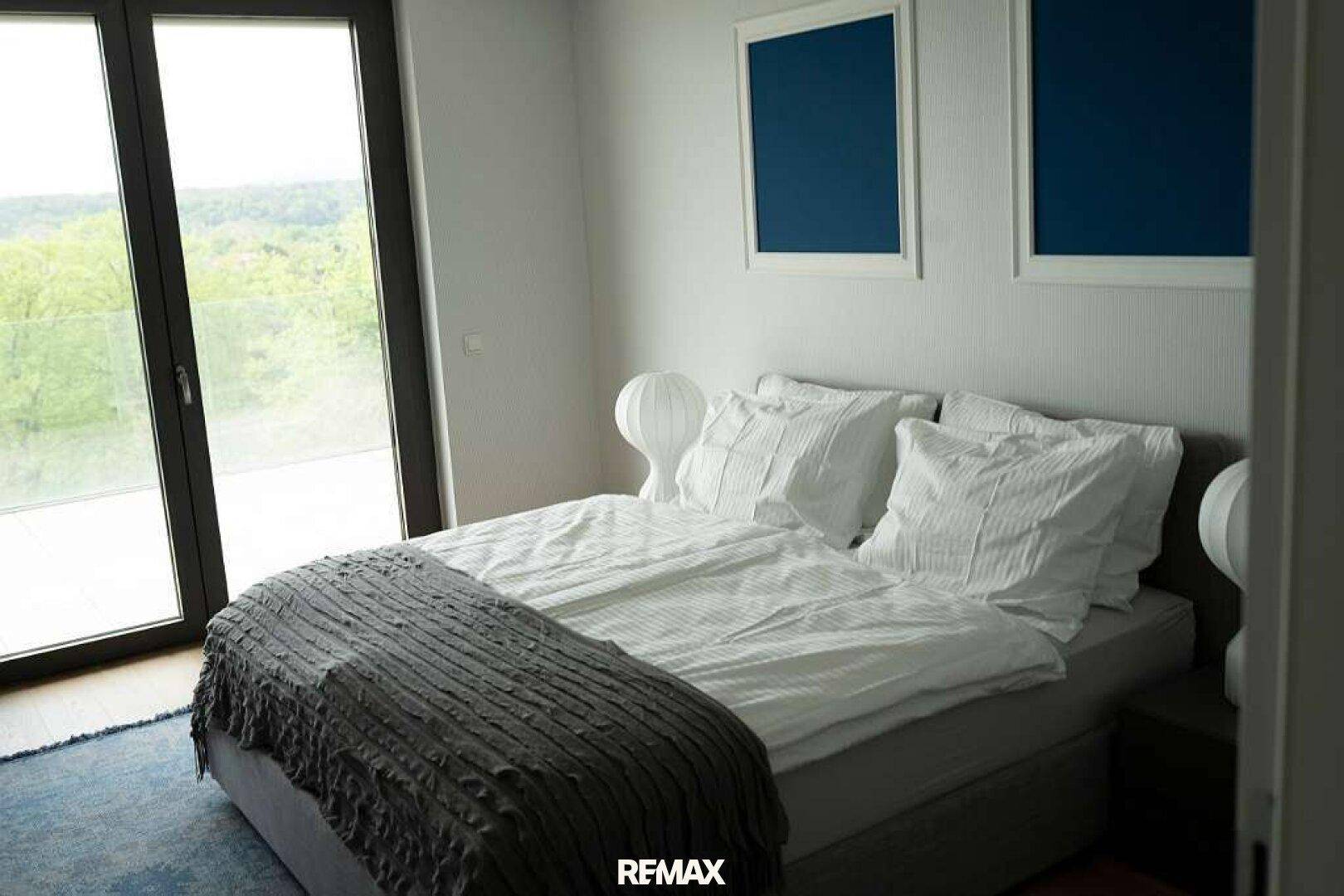 Helles Schlafzimmer mit Doppelbett und großen Glasfenstern, die einen schönen Blick ins Grüne ermöglichen.