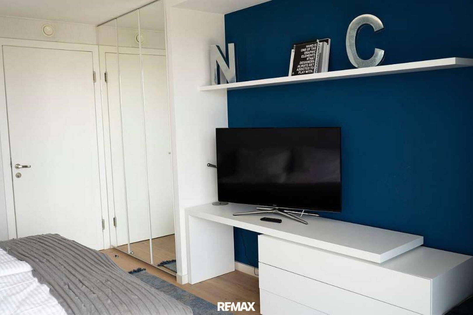 Schlafzimmer mit Fernseher auf weißer Konsole, blauer Akzentwand und verspiegelten Einbauschränken.