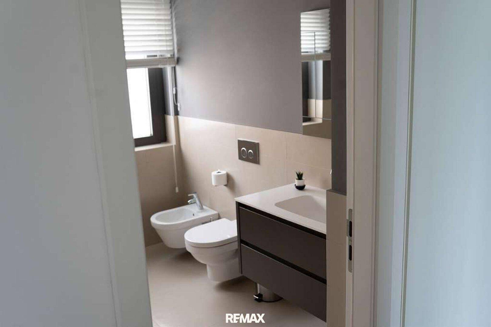 Modernes Badezimmer mit Toilette, Bidet, Einzelwaschbecken und einem Fenster mit Jalousien.