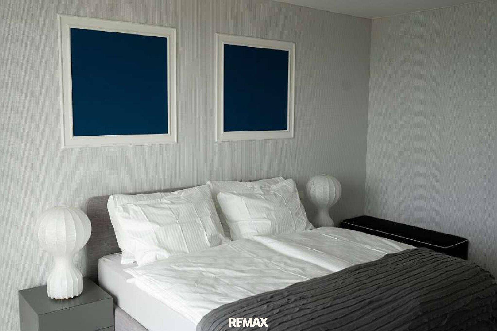 Schlafzimmer mit Doppelbett, weißer Bettwäsche und zwei blauen Bildern an einer hellgrauen Wand.