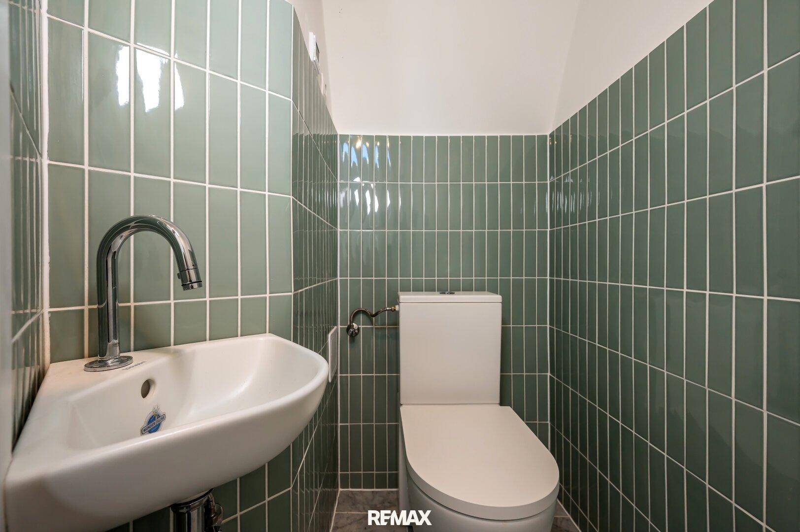 Modern gefliestes Gäste-WC mit grün-glänzenden Wandfliesen und kleinem Waschbecken.