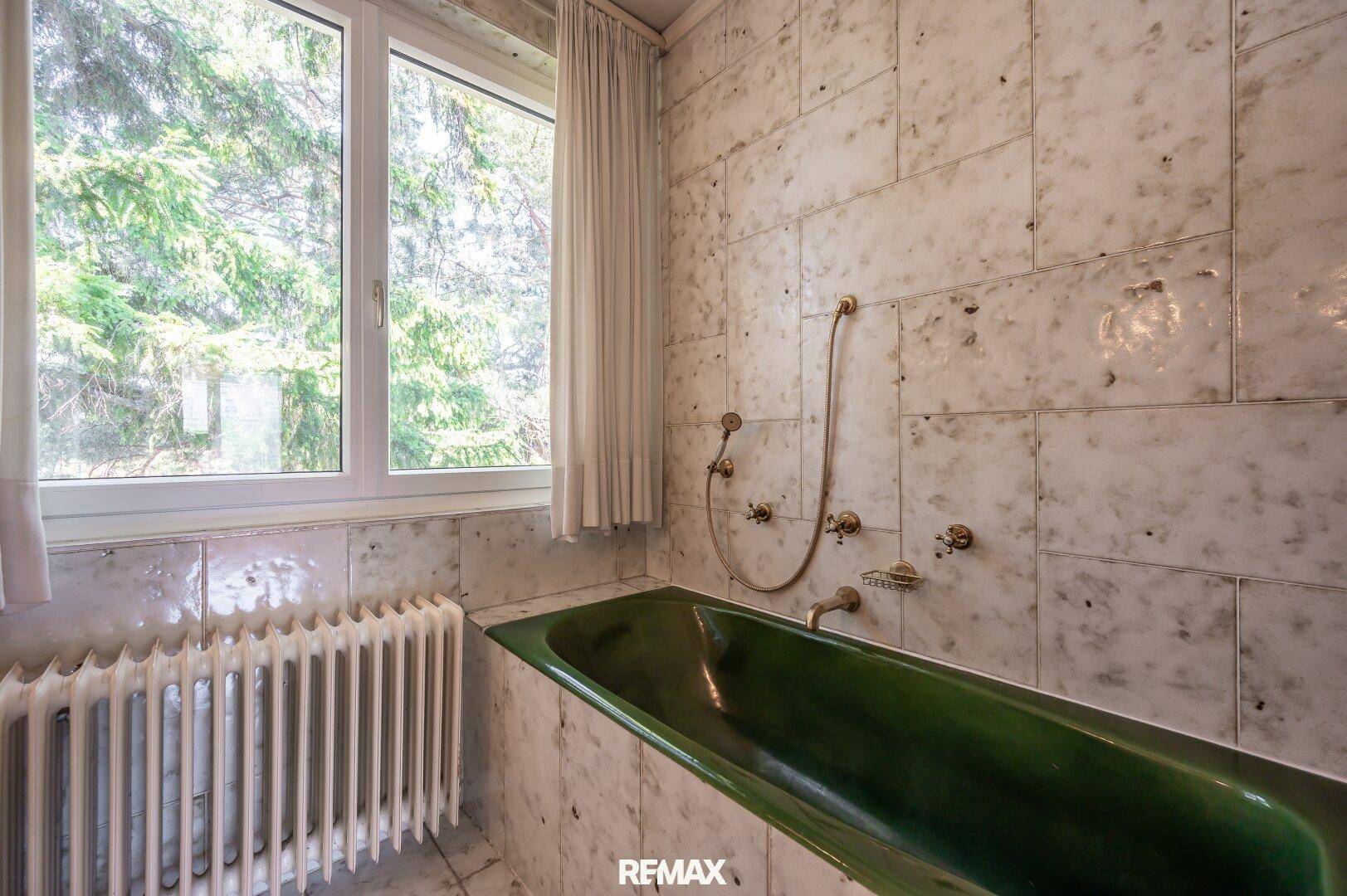 Grüne Badewanne in einem Badezimmer mit Marmorfliesen und Fenster mit Blick ins Grüne.