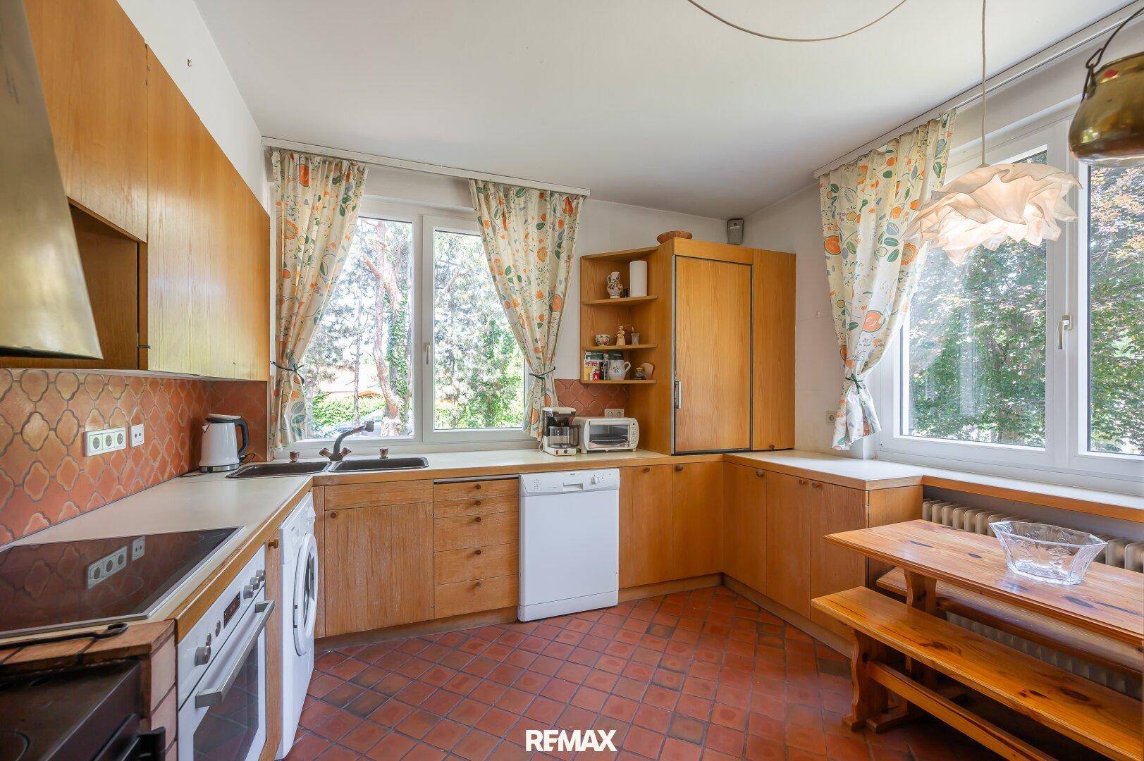 Helle Küche mit Holzeinbauten, Essbereich und großen Fenstern mit Blick ins Grüne.