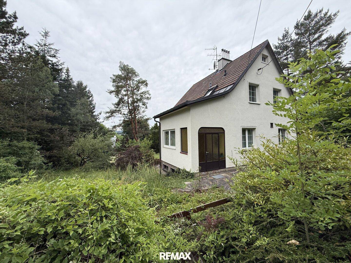 Älteres Einfamilienhaus mit Giebeldach und verwildertem Garten, umgeben von hohen Bäumen.