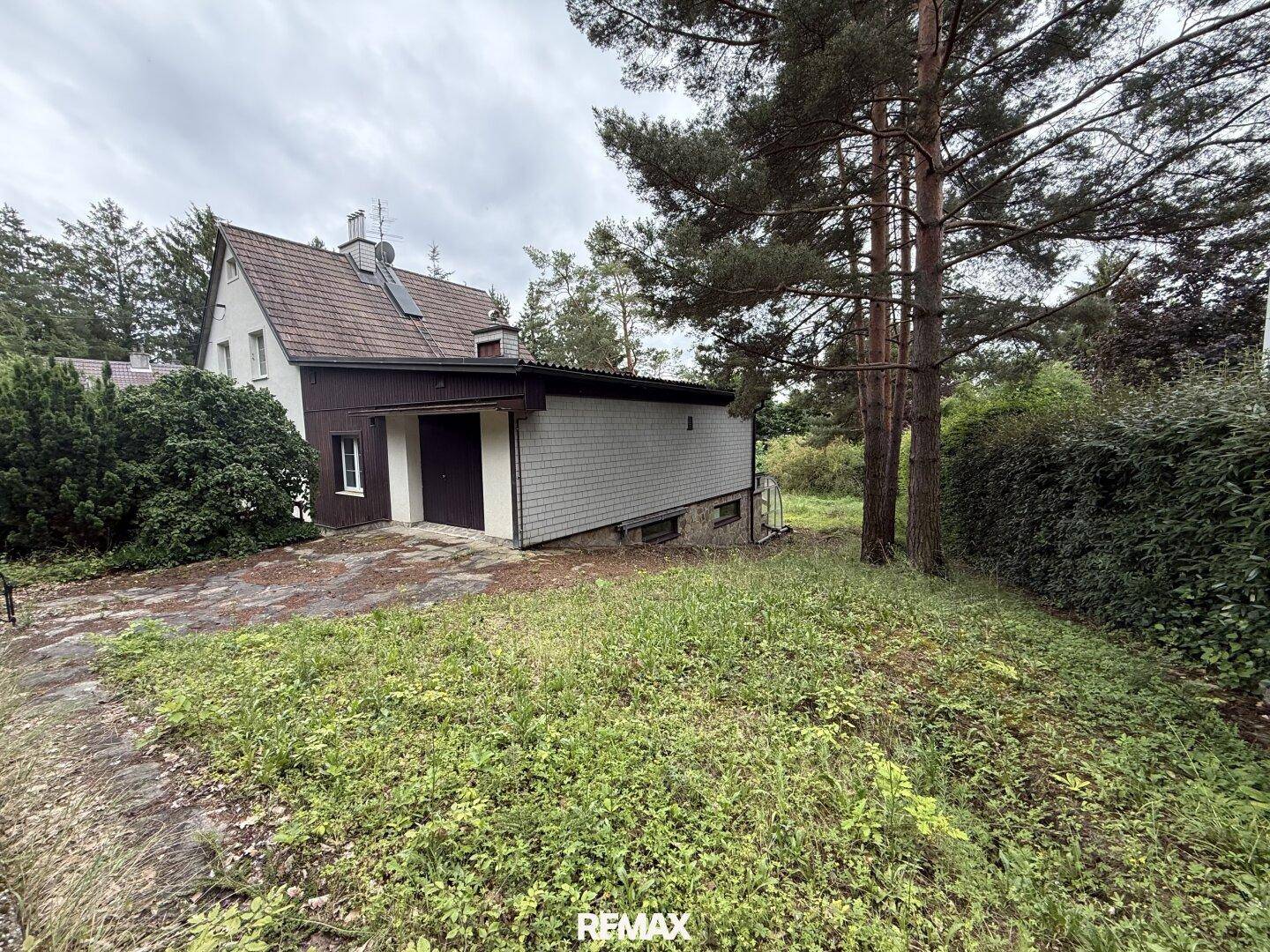 Einfamilienhaus mit Anbau und verwildertem Vorgarten, umgeben von hohen Bäumen.