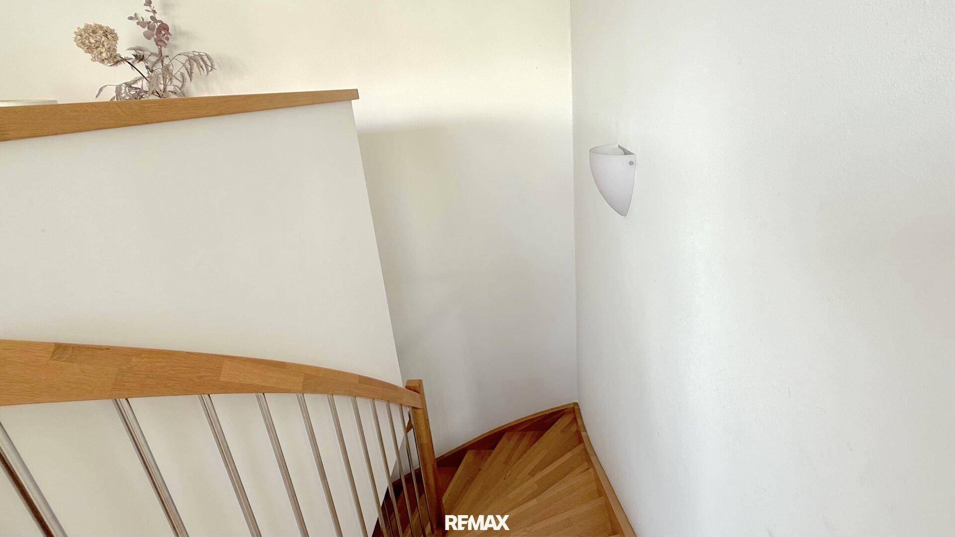 Holztreppe mit Metallgeländer führt in den oberen Bereich, helle Wände und indirekte Beleuchtung.