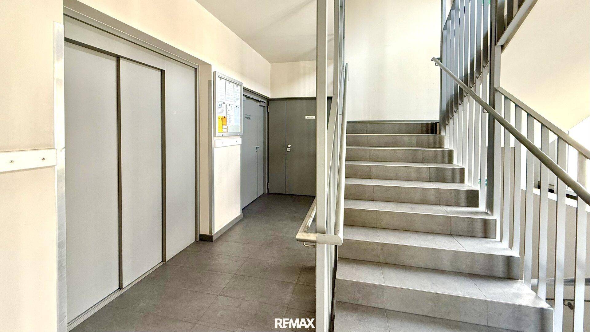 Treppenhaus und Aufzug in einem modernen Gebäude mit Fliesenboden, der den Zugang zu den Etagen ermöglicht.