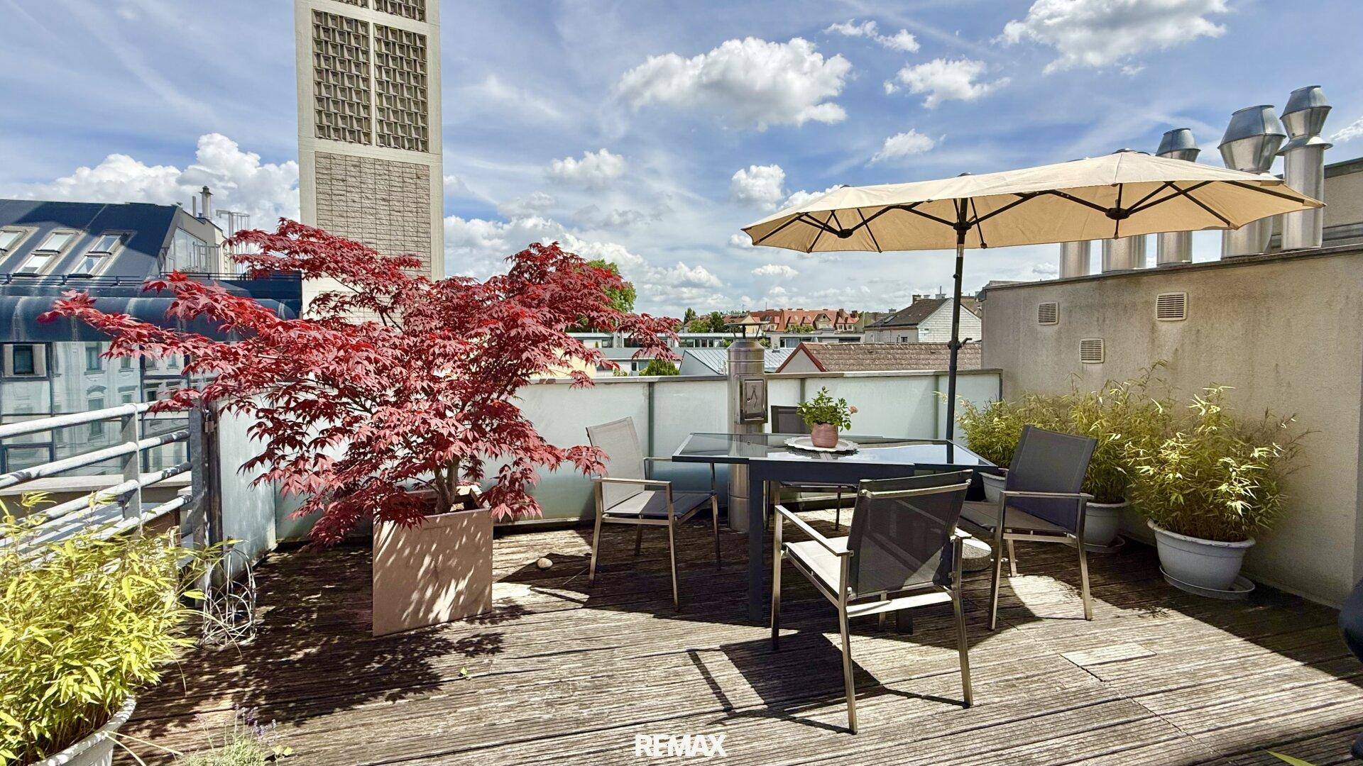 Geräumige Dachterrasse mit Holzboden, Sitzgelegenheiten und einem Sonnenschirm unter blauem Himmel.
