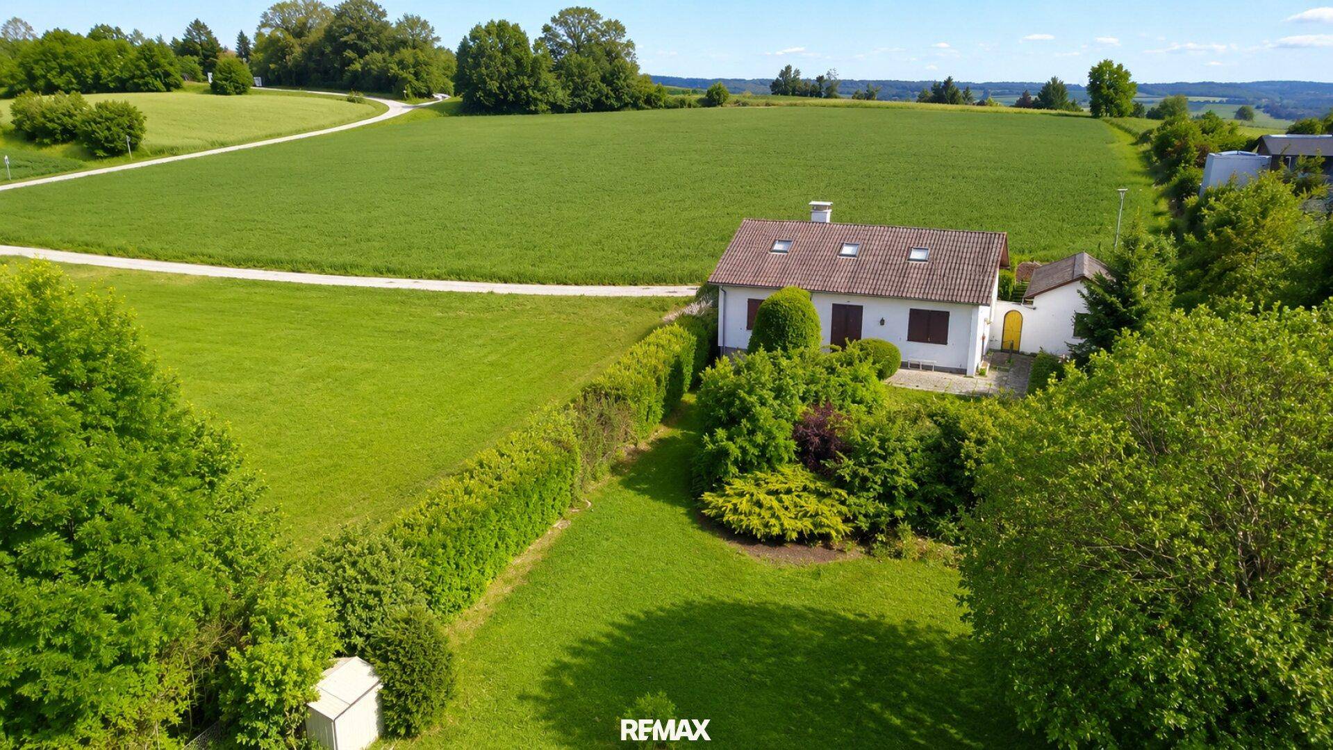 Haus in ländlicher Umgebung, umgeben von weiten grünen Feldern unter blauem Himmel.