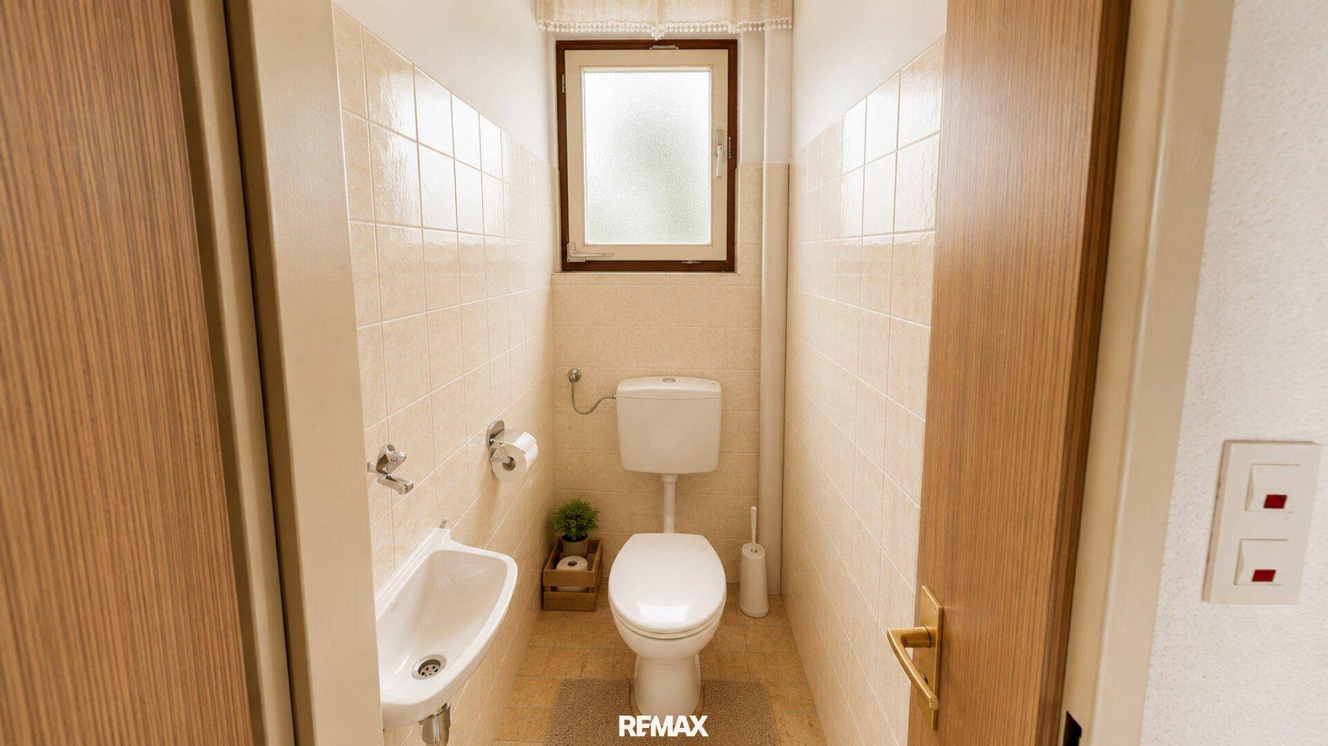 Separates WC mit Toilette, kleinem Waschbecken und Fenster, in hellen Fliesen gehalten.