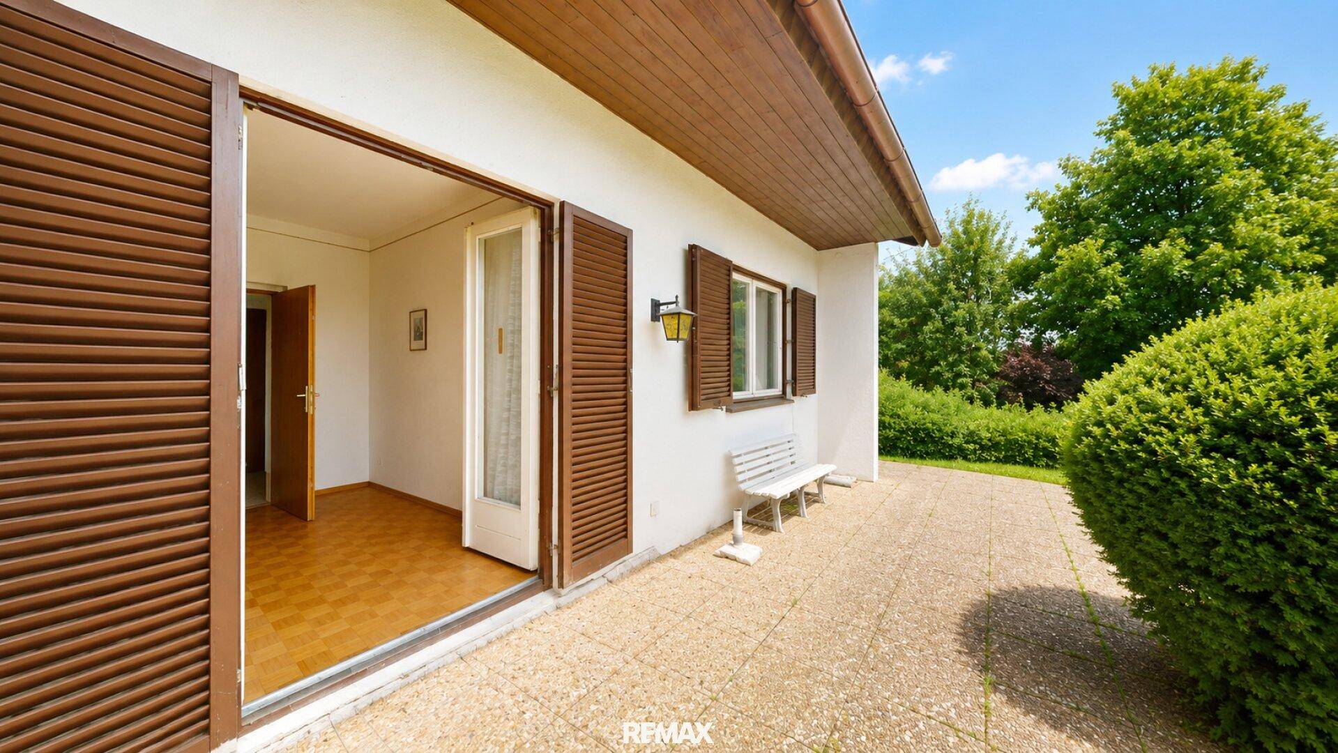 Terrasse mit Zugang zum Innenbereich, umgeben von grünen Sträuchern und mit Holzboden.
