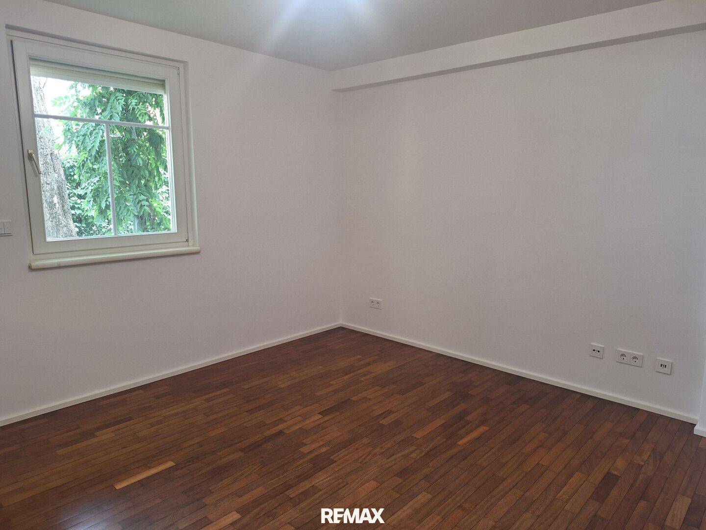 Helles Schlafzimmer mit Parkettboden und einem Fenster mit Blick ins Grüne.