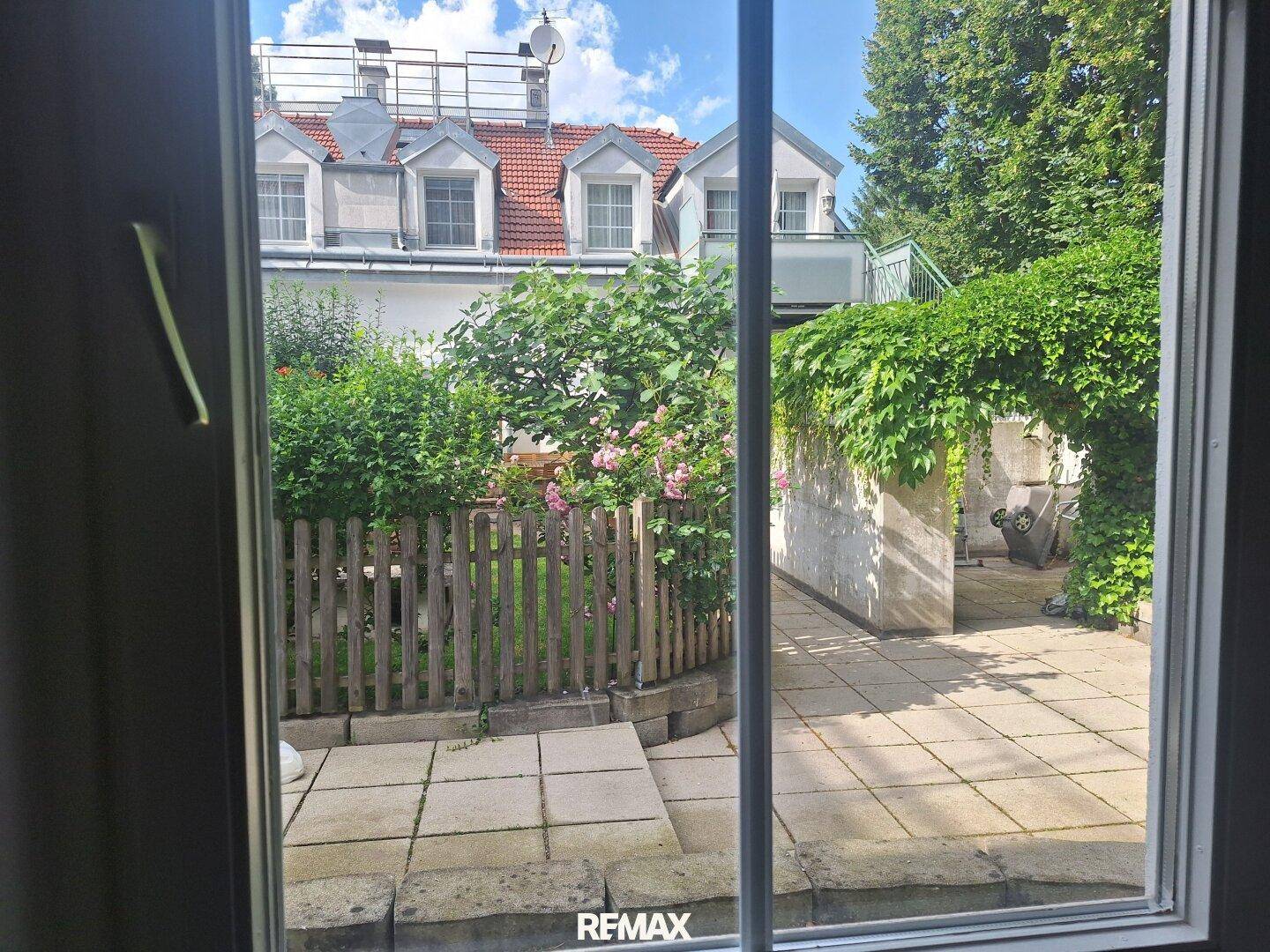 Aussicht aus dem Fenster auf eine gepflasterte Terrasse mit üppigem Grün und einem sonnigen Bereich.