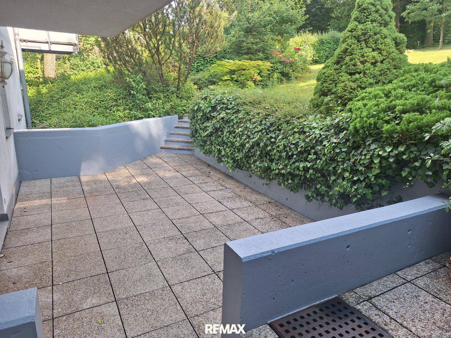 Sonniger Terrassenbereich mit Steinplatten und einer grünen Hecke, ideal zum Entspannen im Freien.