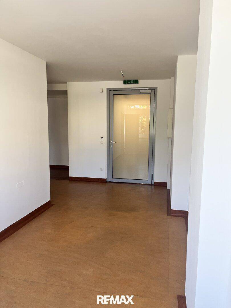 Heller Flur mit dunklem Bodenbelag und einer modernen Glastür, die den Zugang zum Bereich ermöglicht.