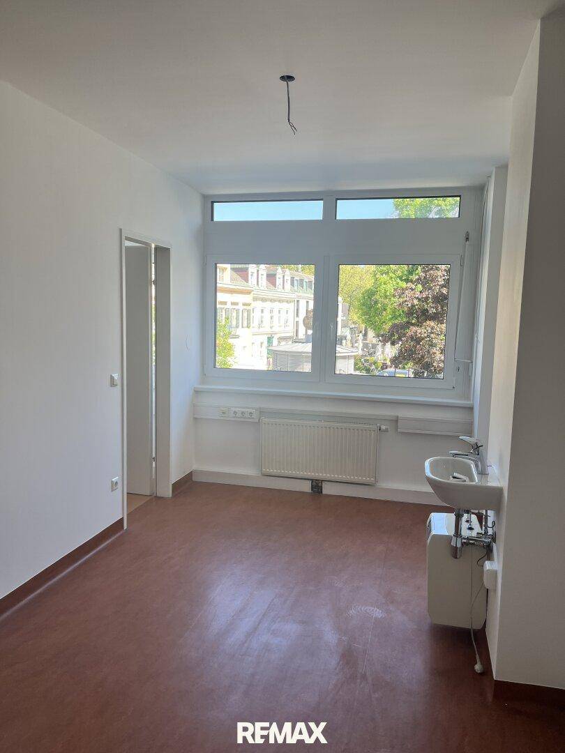 Heller Raum mit Fenster, dunklem Bodenbelag und einem Waschbecken, geeignet als Büro oder Behandlungszimmer.