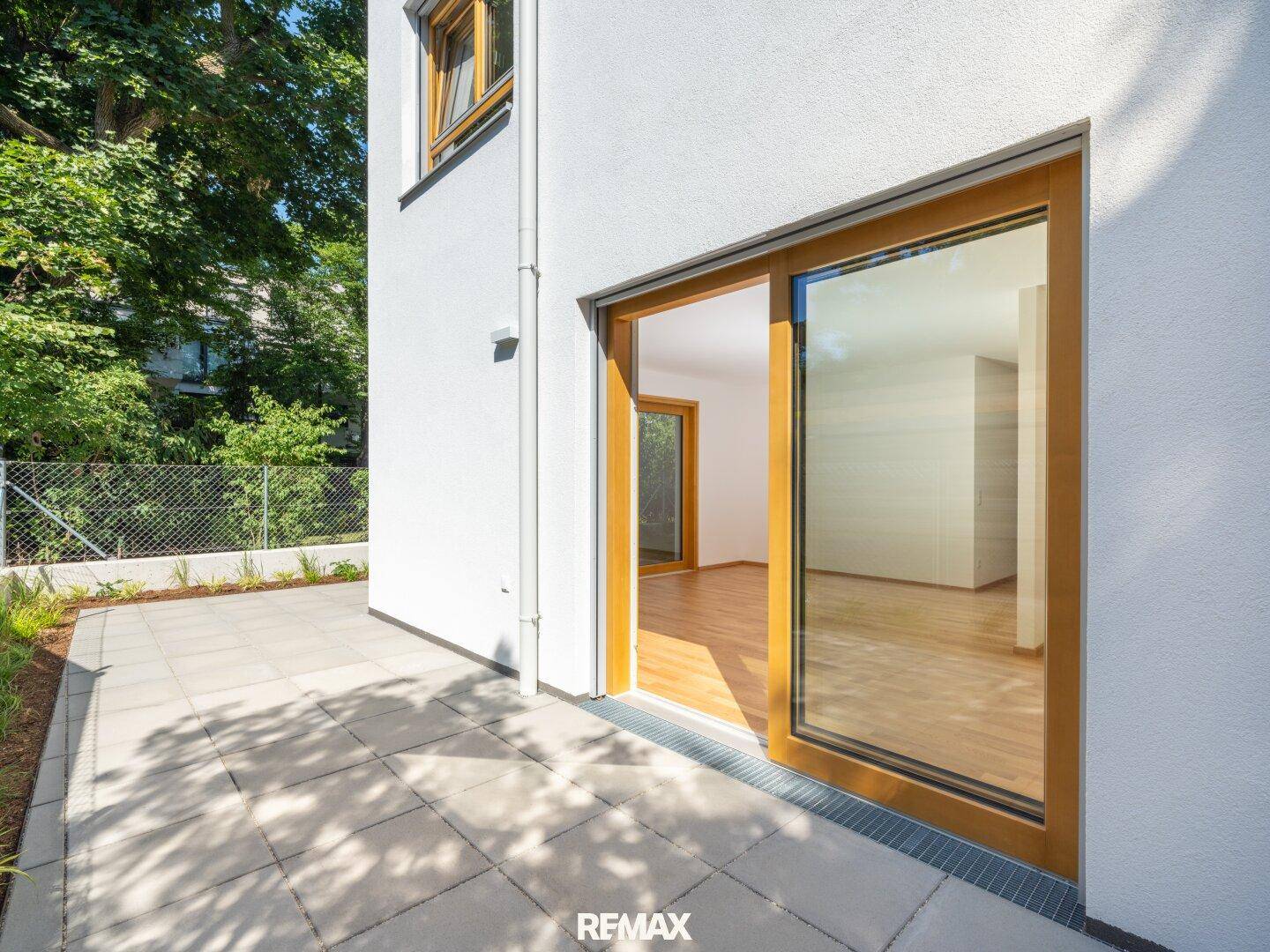 Moderne Gebäudefassade mit großer Glasschiebetür, die zu einer sonnigen, gepflasterten Terrasse führt.