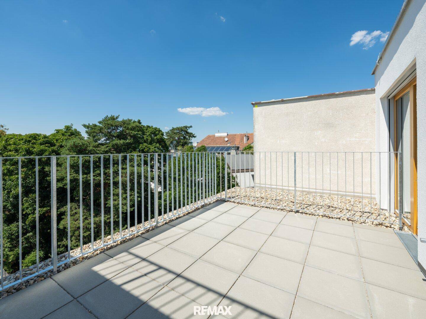 Großer Balkon mit Fliesenboden und Blick auf grüne Bäume unter blauem Himmel.