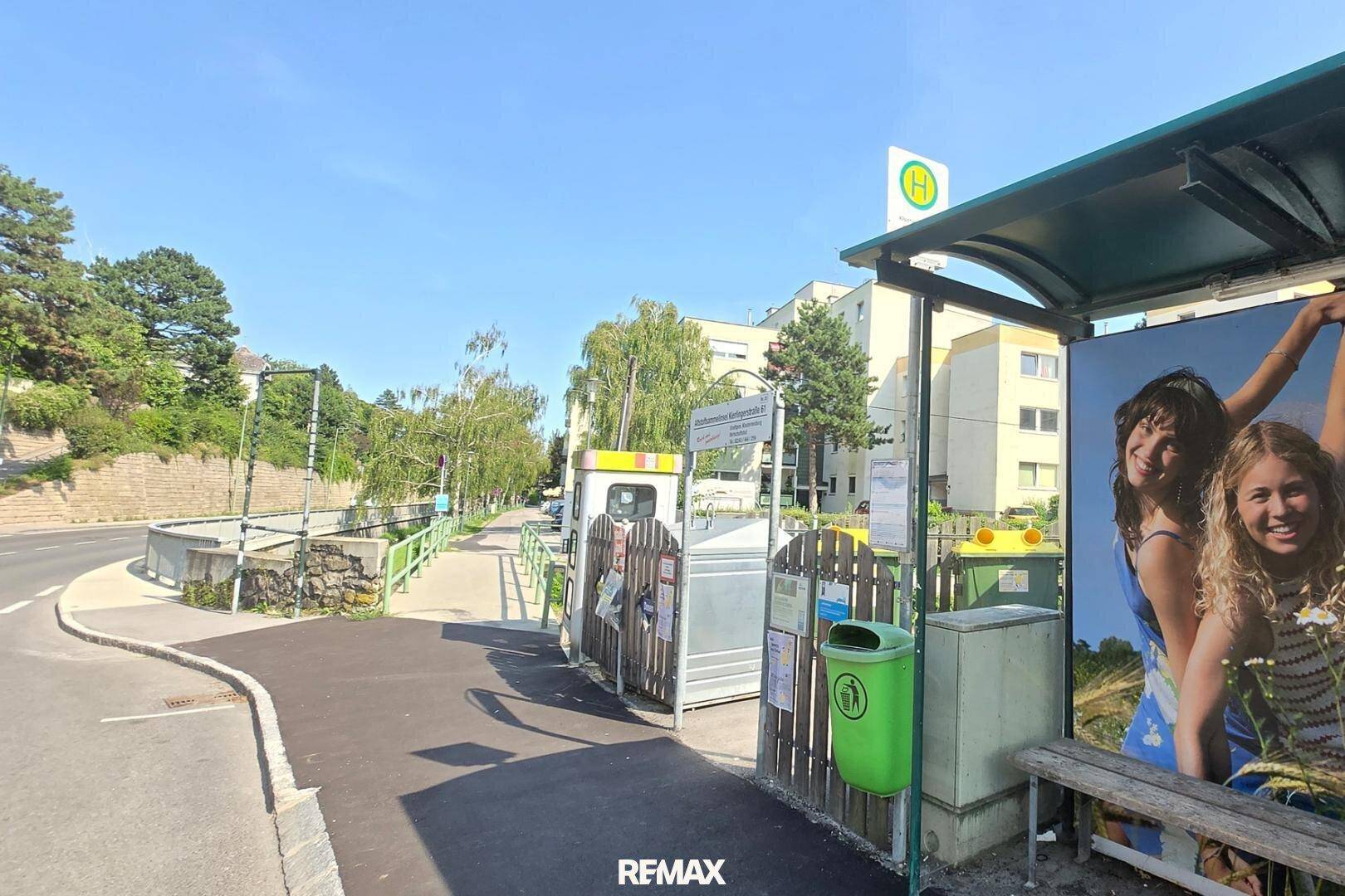 Straßenansicht mit Bushaltestelle, Recyclingcontainern und Wohngebäuden im Hintergrund.