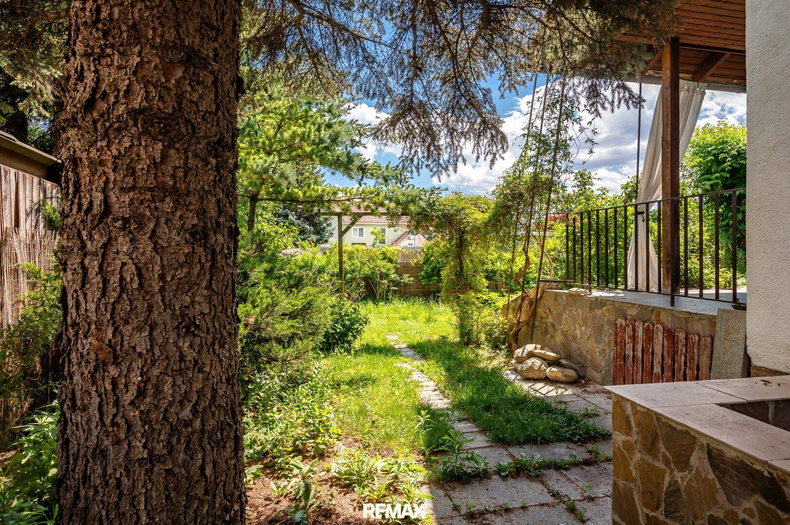 Grüner Gartenbereich mit altem Baum und Zugang zu einer überdachten Terrasse.