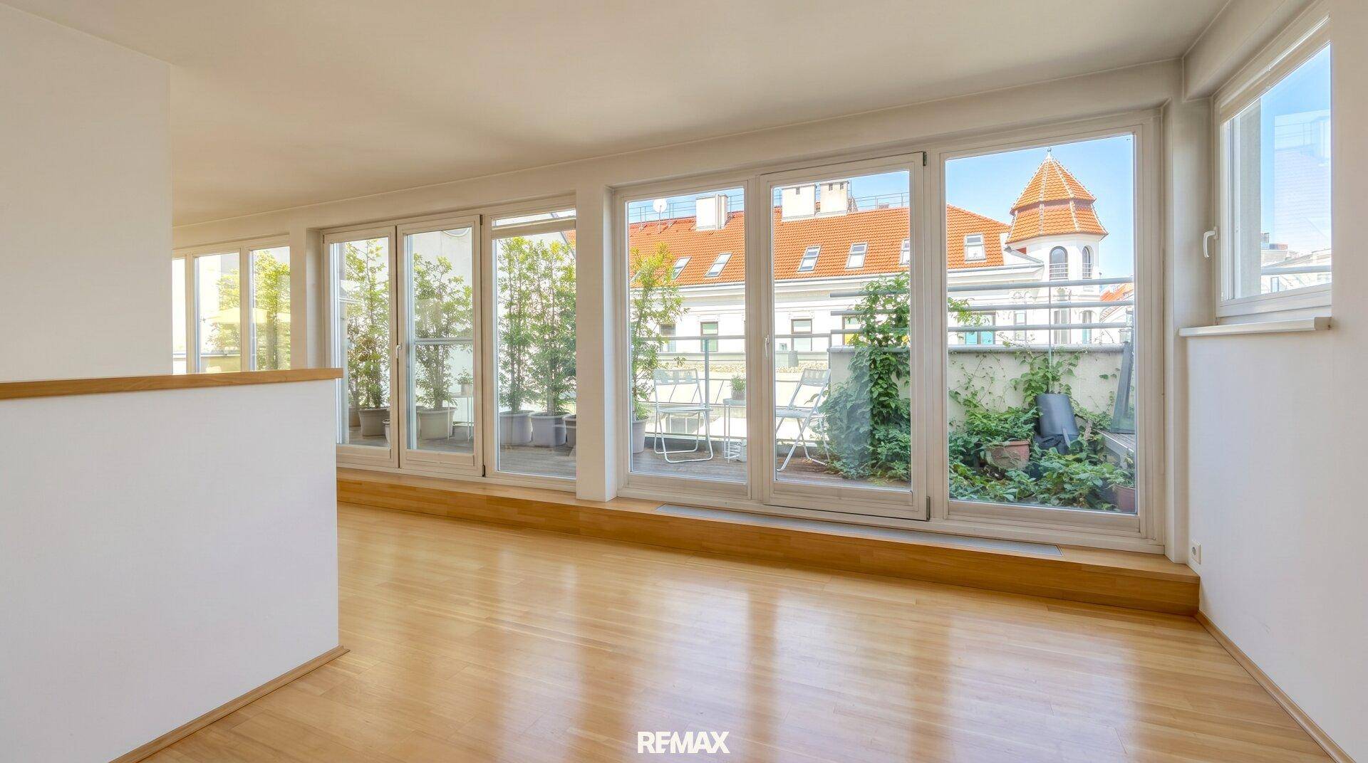 Lichtdurchfluteter Wohnbereich mit Holzboden und Blick auf eine begrünte Terrasse und historische Gebäude.