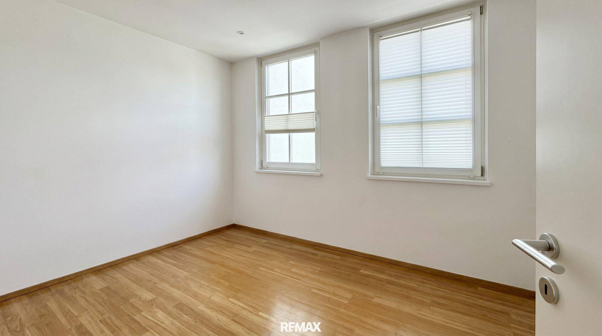 Helles Schlafzimmer mit zwei Fenstern, Holzboden und weißen Wänden, ideal für Ruhe und Entspannung.