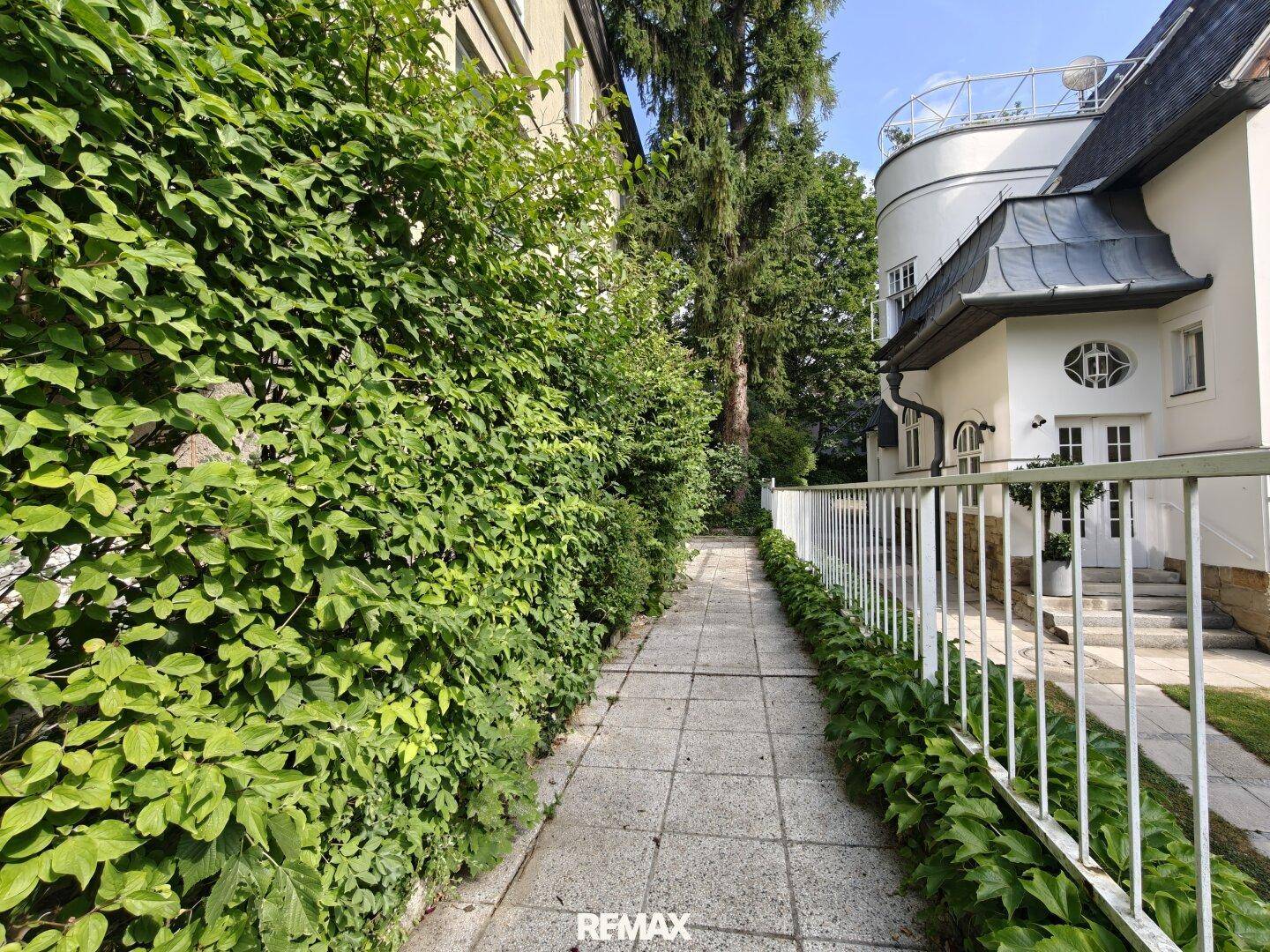 Grüner Weg entlang einer Hausfassade mit üppiger Bepflanzung und einem weißen Zaun.