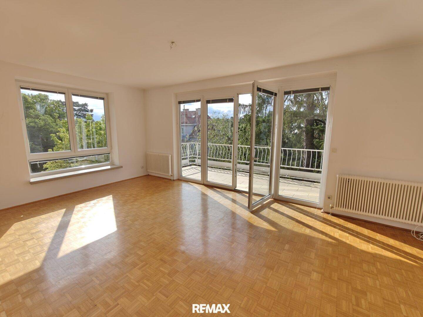 Geräumiges, leeres Wohnzimmer mit Parkettboden und großen Fenstern, die viel Licht hereinlassen.