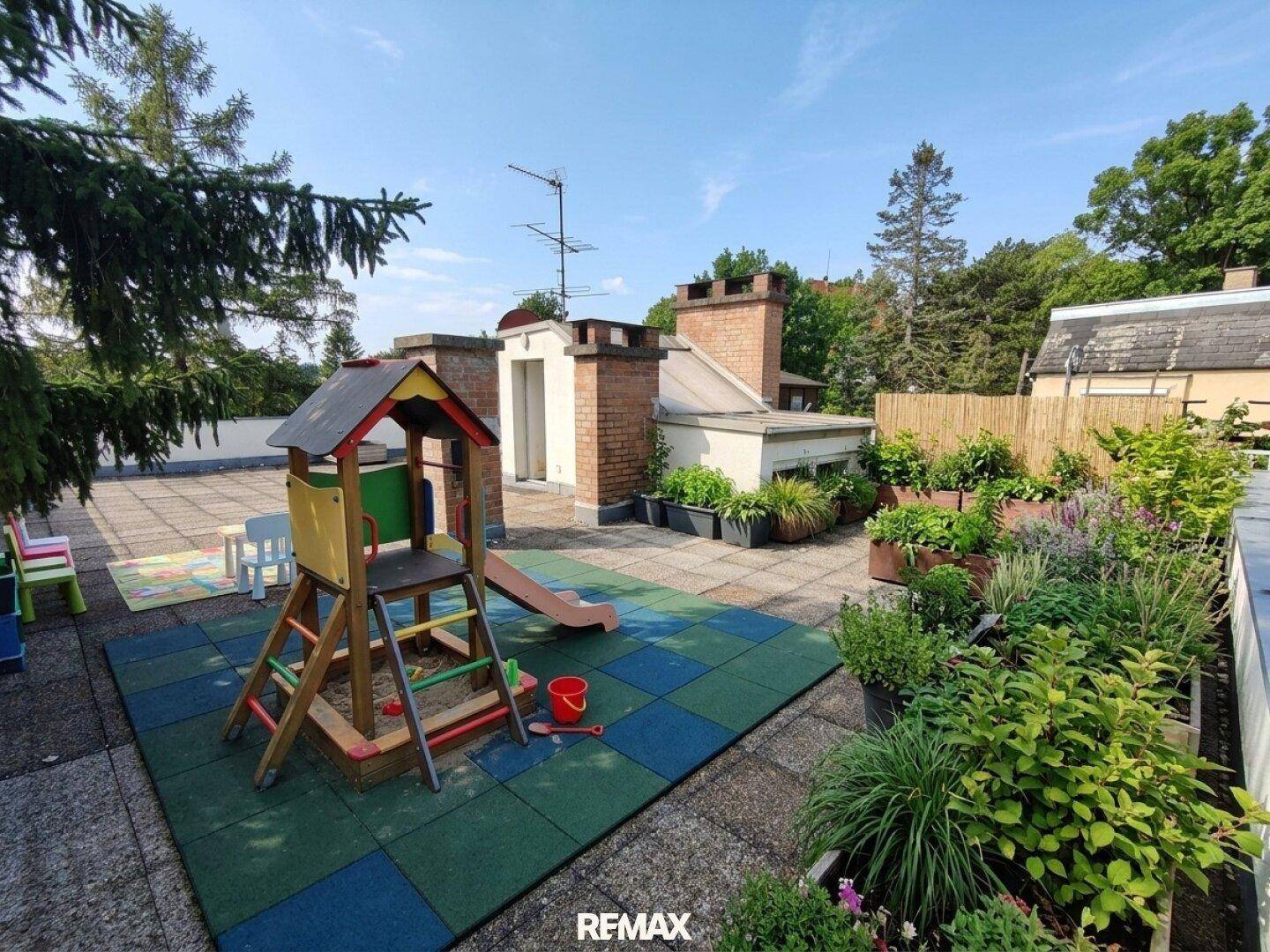 Dachterrasse mit Kinderspielplatz und bepflanzten Hochbeeten, umgeben von Bäumen.