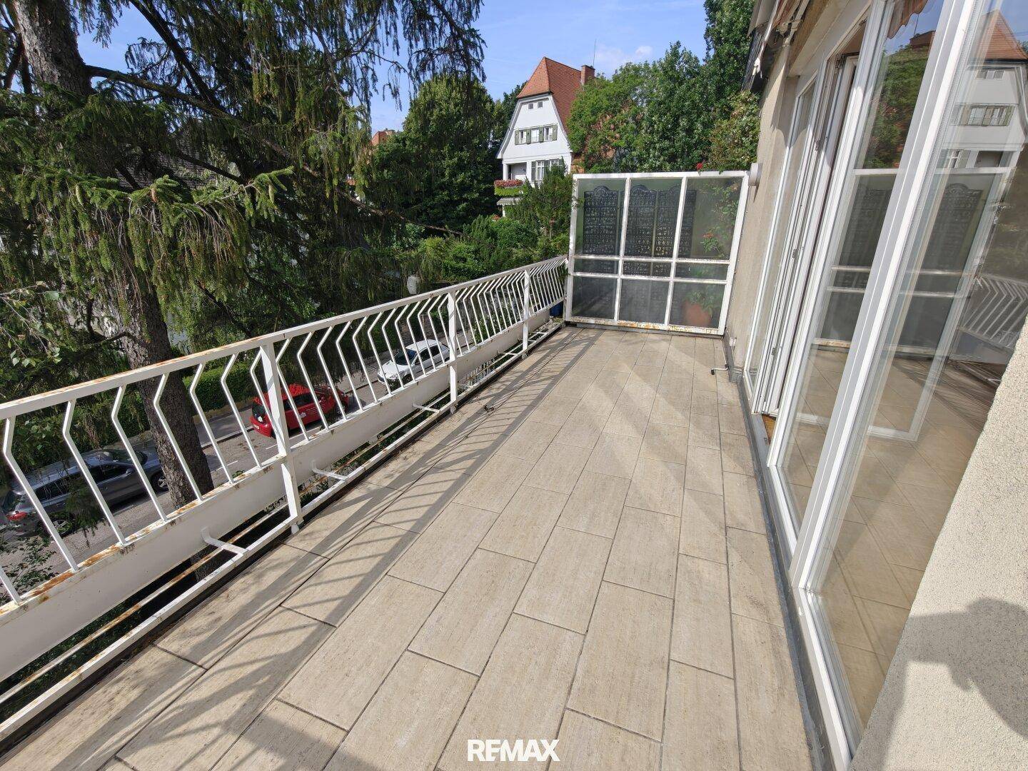 Langer, schmaler Balkon mit Fliesenboden und weißem Geländer, bietet Blick auf die Straße und Bäume.