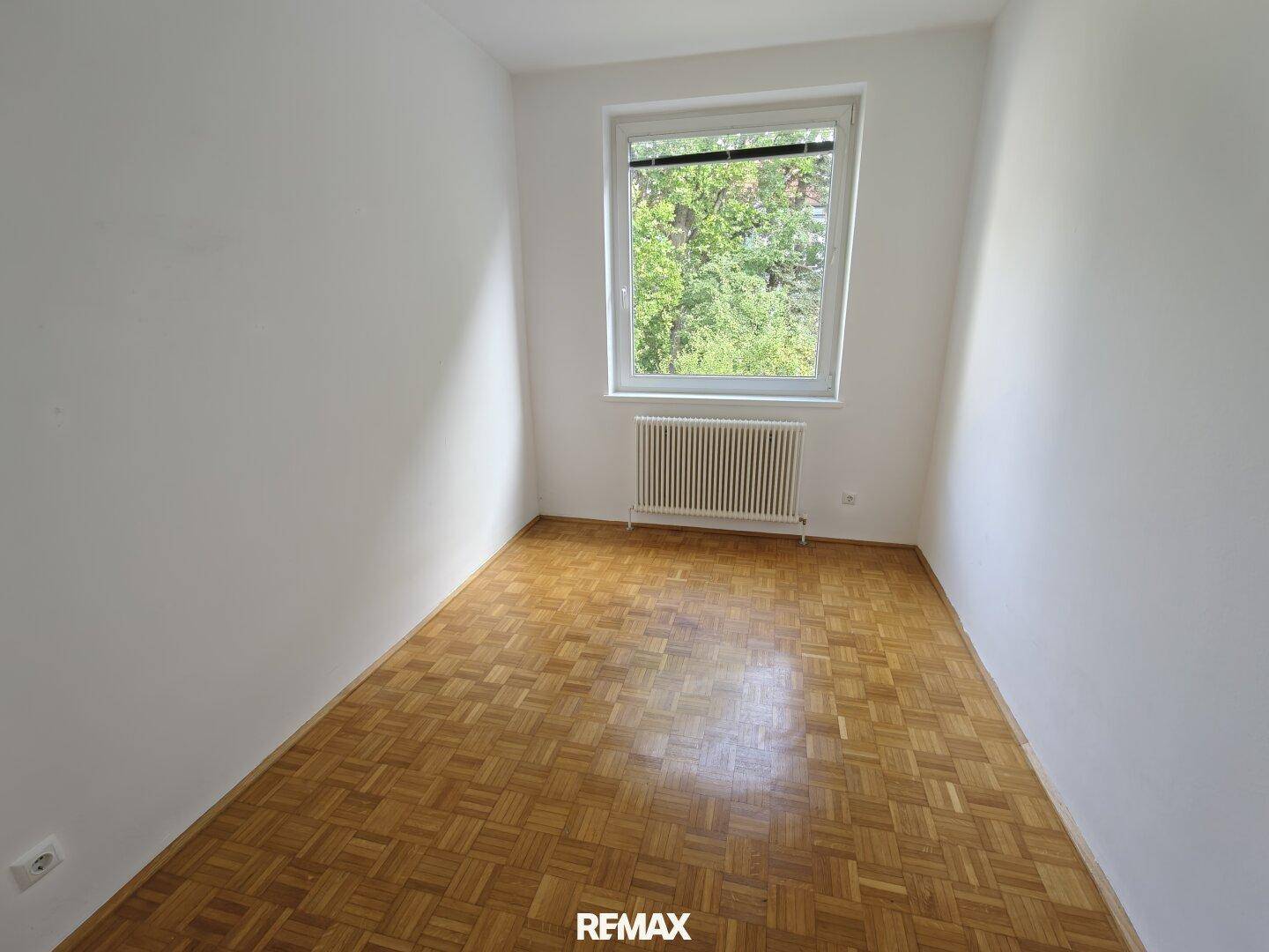 Leeres Schlafzimmer mit Parkettboden und einem großen Fenster, das viel Tageslicht hereinlässt.