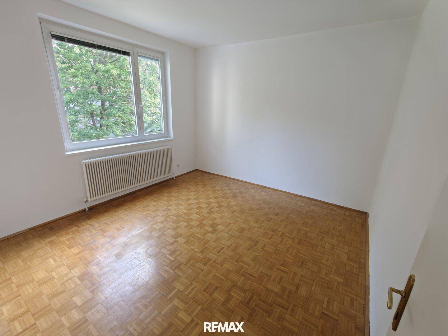 Leeres Schlafzimmer mit Parkettboden und einem großen Fenster, das viel Tageslicht hereinlässt.