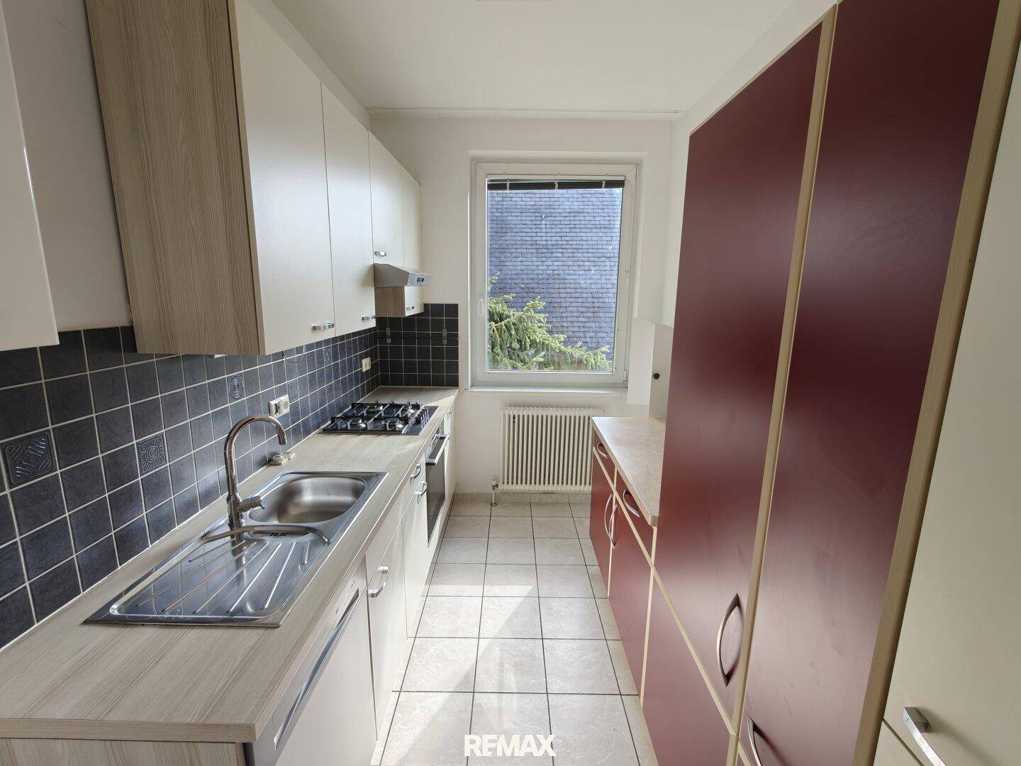 Schmale Küche mit Gasherd, Fliesenboden und Fenster, das Tageslicht hereinlässt.