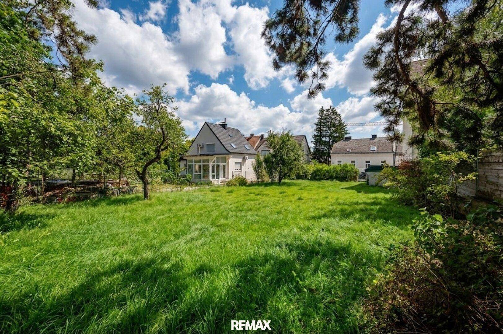 Große Rasenfläche mit altem Baumbestand und einem Wohnhaus im Hintergrund unter blauem Himmel.