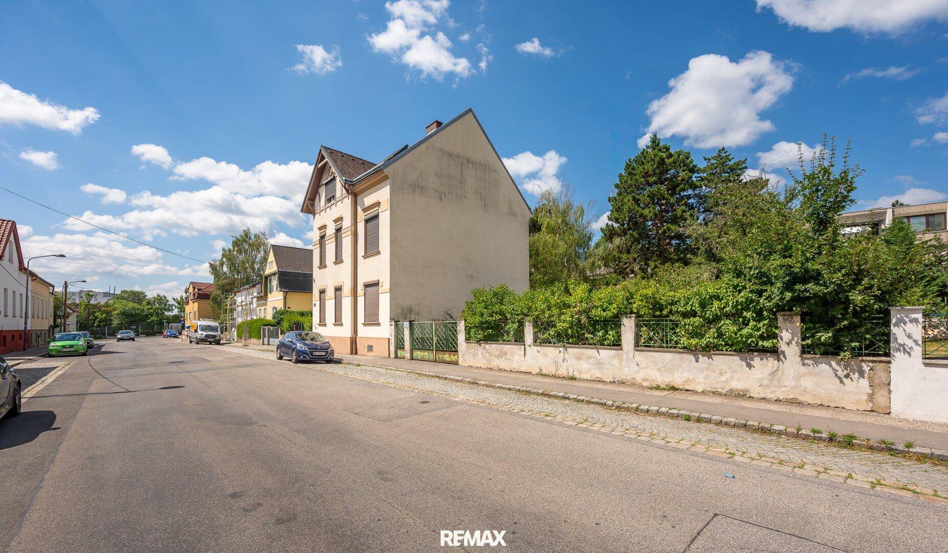 Blick auf die Fassade eines Gebäudes von der Straße aus, umgeben von Bäumen und Nachbarhäusern.