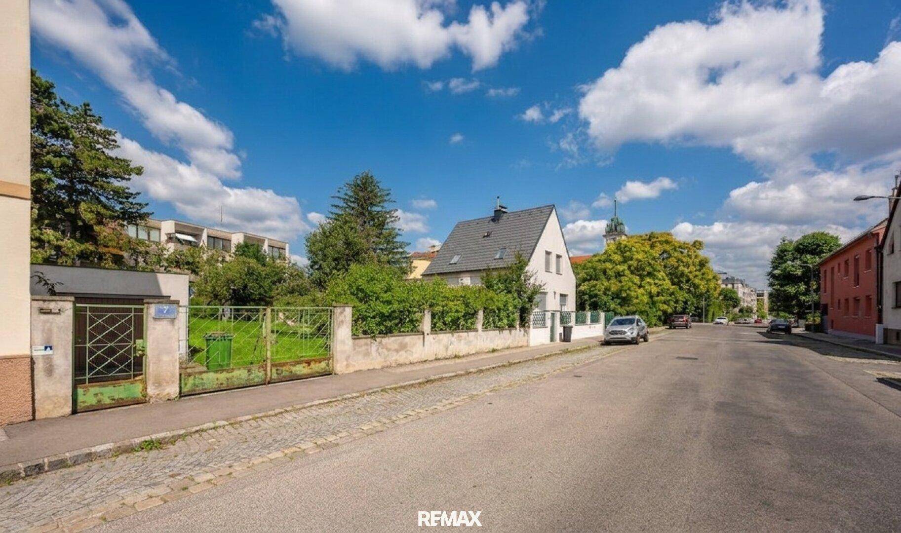 Straßenansicht des Grundstücks mit Zaun, Tor und umliegenden Gebäuden unter blauem Himmel.
