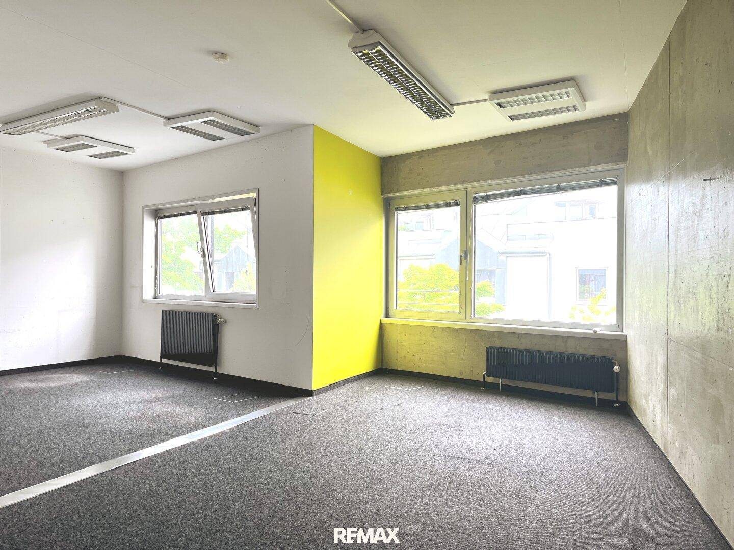 Geräumiger Büroraum mit grauem Teppichboden, Sichtbetonwand und einem gelben Akzent.