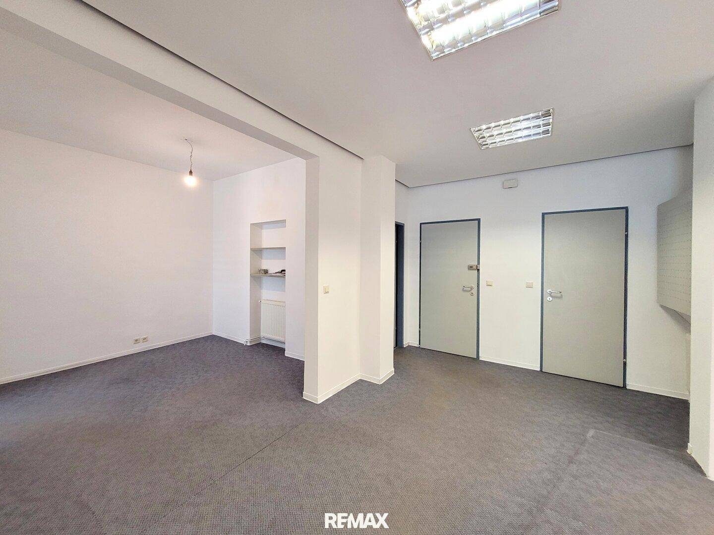 Geräumiges Büro mit grauem Teppichboden, weißen Wänden, zwei Türen und einer Nische mit Regalen.