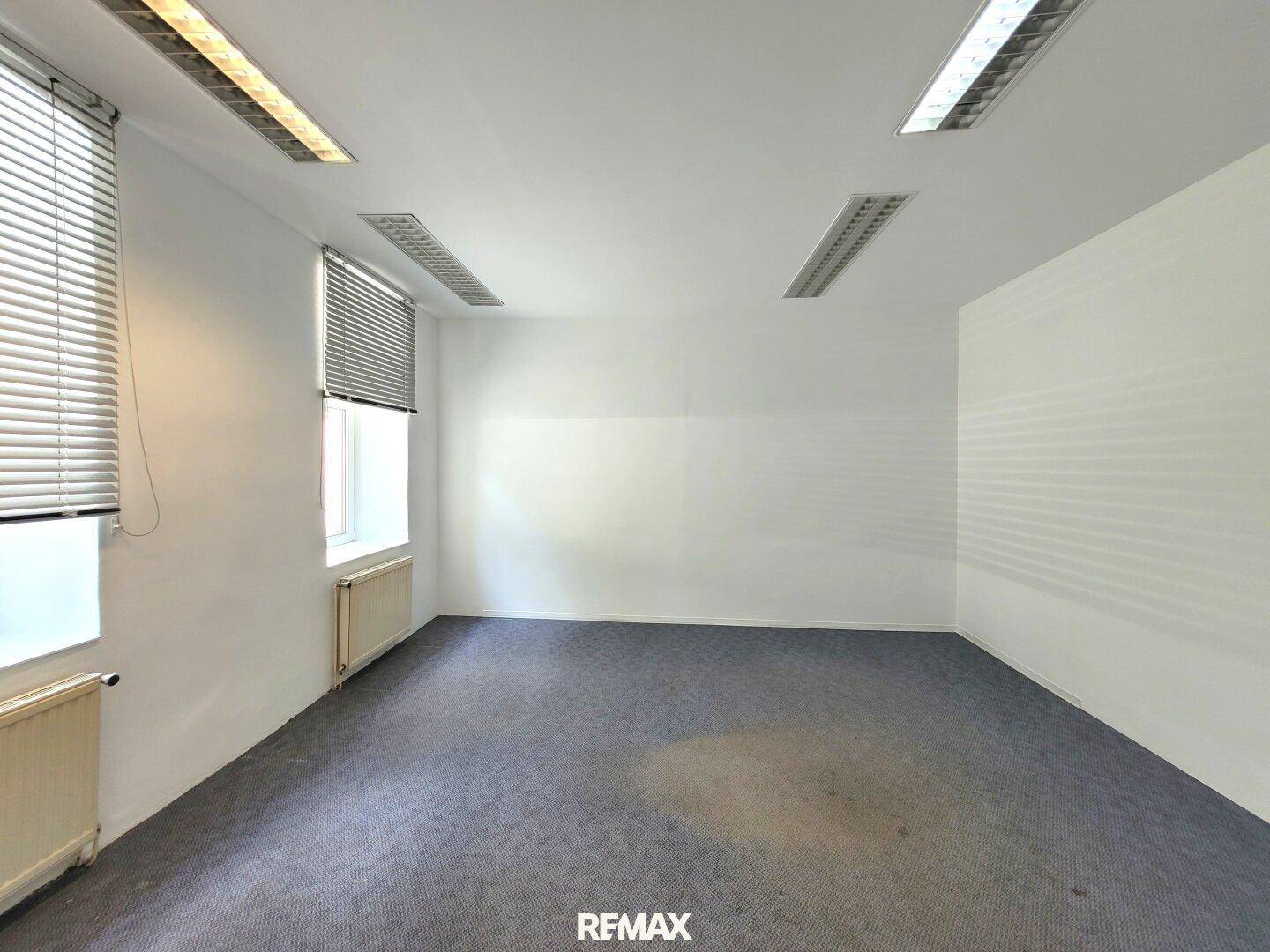 Helles Büro mit grauem Teppichboden, weißen Wänden, Fenstern mit Jalousien und Heizkörpern.