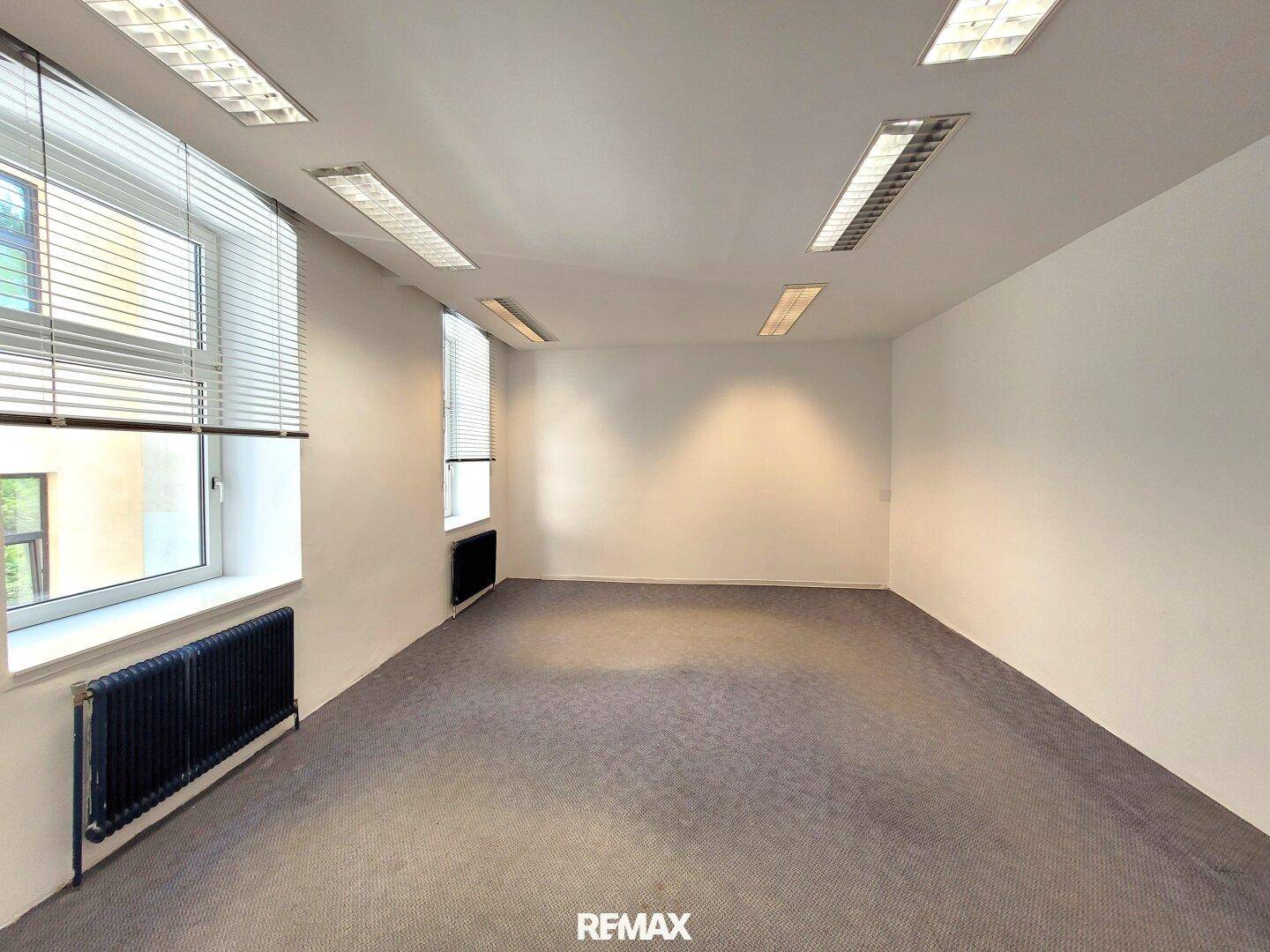 Leeres Büro mit grauem Teppichboden, weißen Wänden, Fenstern mit Jalousien und schwarzen Heizkörpern.