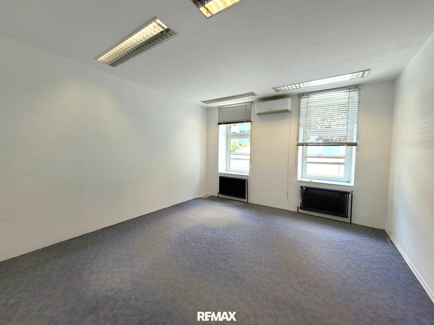 Bürofläche mit grauem Teppichboden, weißen Wänden, Fenstern, Heizkörpern und einer Klimaanlage.