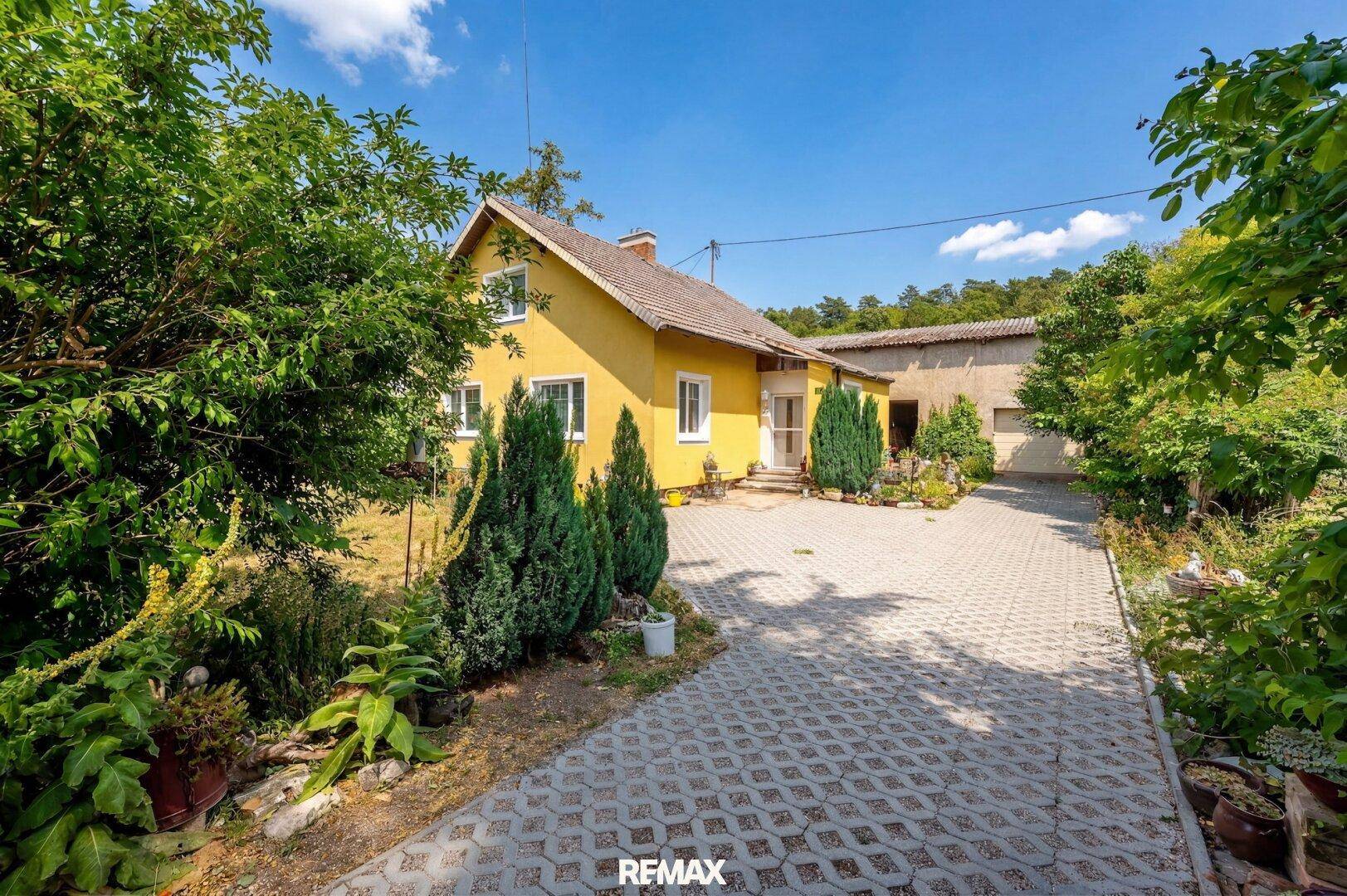 Gelbes Einfamilienhaus mit gepflasterter Auffahrt und umgebender grüner Vegetation unter blauem Himmel.