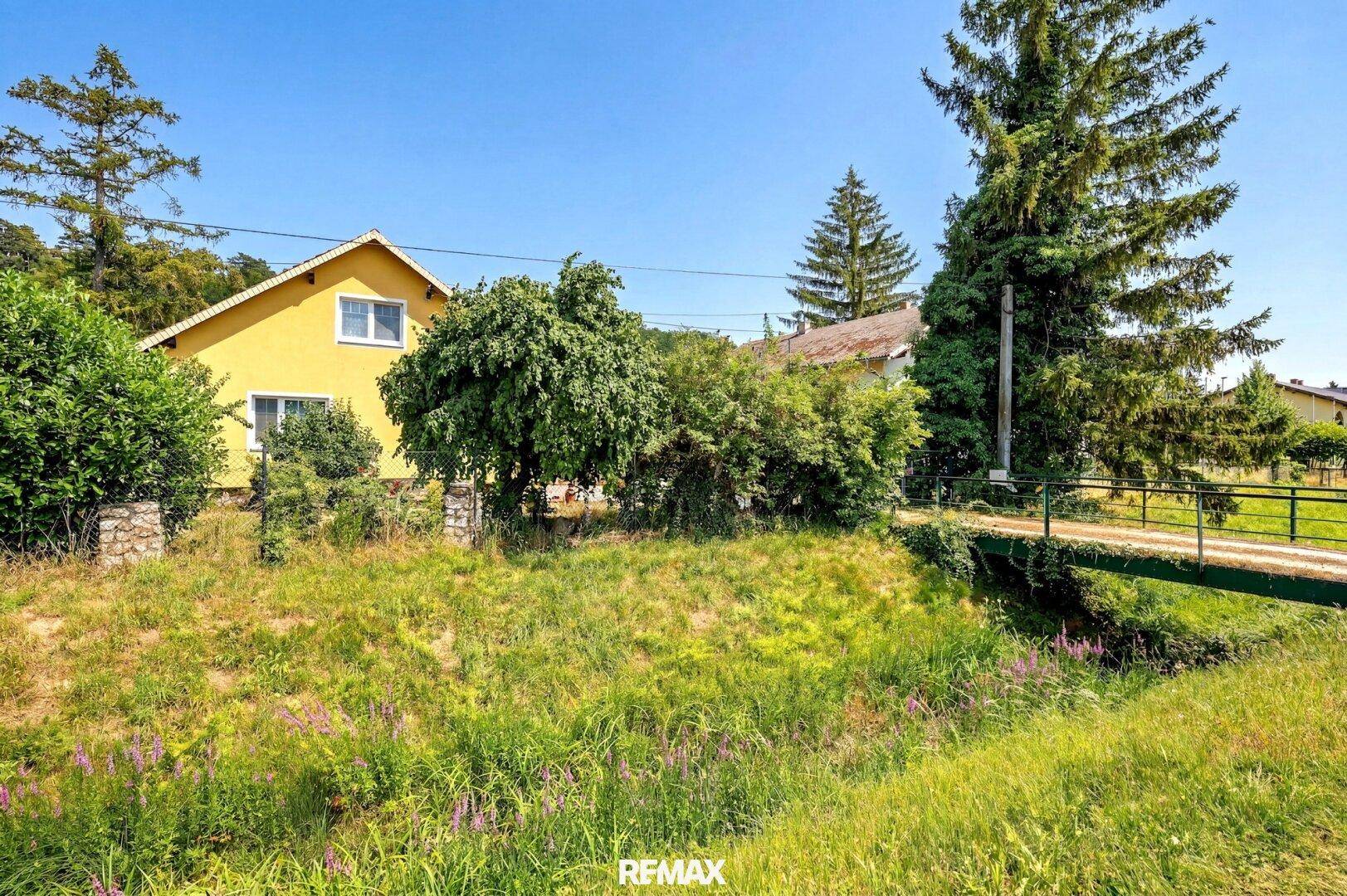 Gelbes Haus in ländlicher Umgebung mit einem kleinen Bach und einer Brücke im Vordergrund.