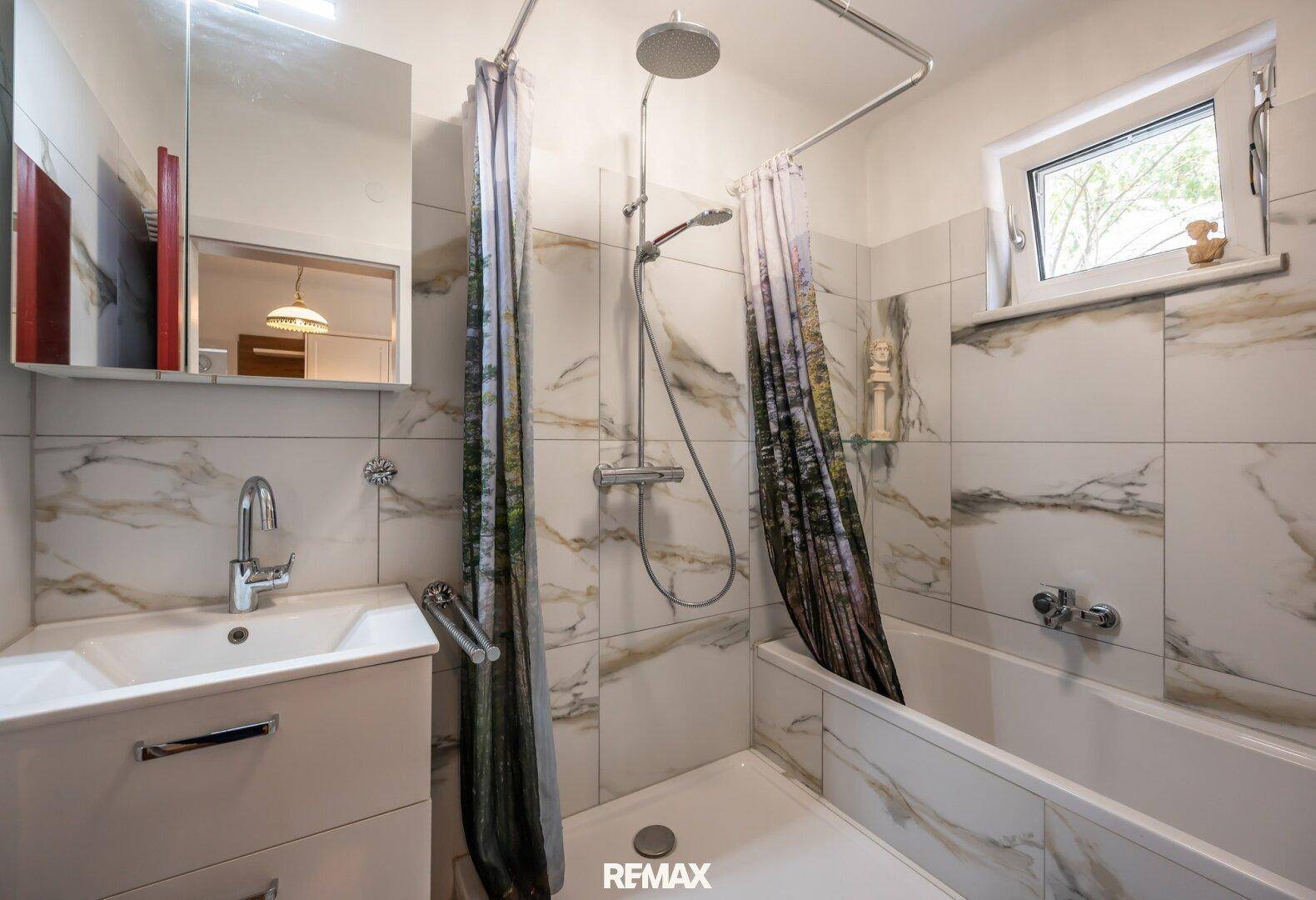 Helles Badezimmer mit Badewanne, Dusche und einem Fenster, gefliest in Marmoroptik.
