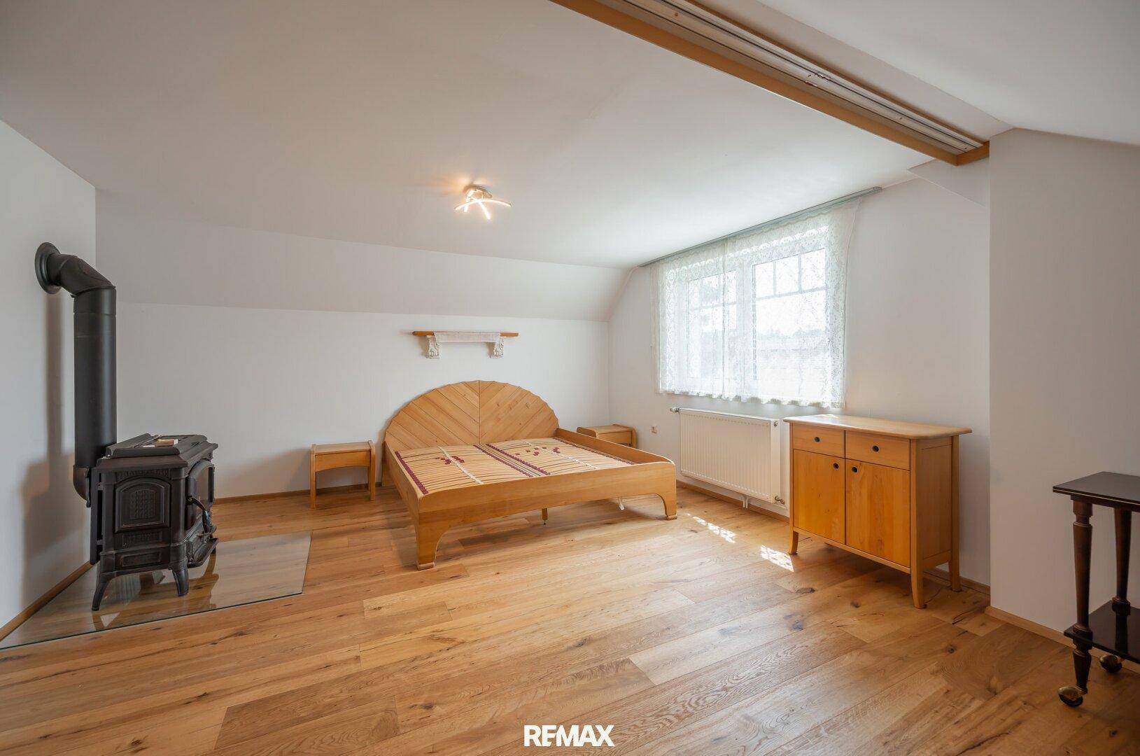 Geräumiges Schlafzimmer im Dachgeschoss mit Holzboden, Holzofen und einem großen Fenster.