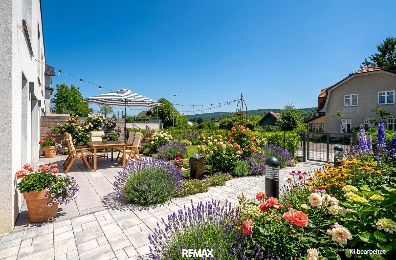 Einladende Terrasse mit Esstisch und Sonnenschirm, umgeben von einem blühenden Garten unter blauem Himmel.