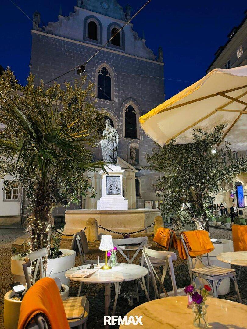 Beleuchteter Außenbereich eines Restaurants bei Nacht mit historischem Gebäude und Statue im Hintergrund.