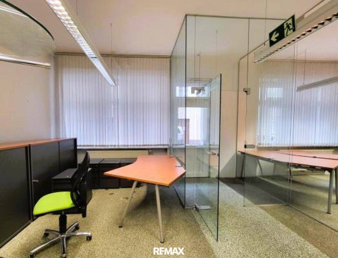 Büroraum mit Glasabtrennung, Schreibtisch und Fenster mit Jalousien.