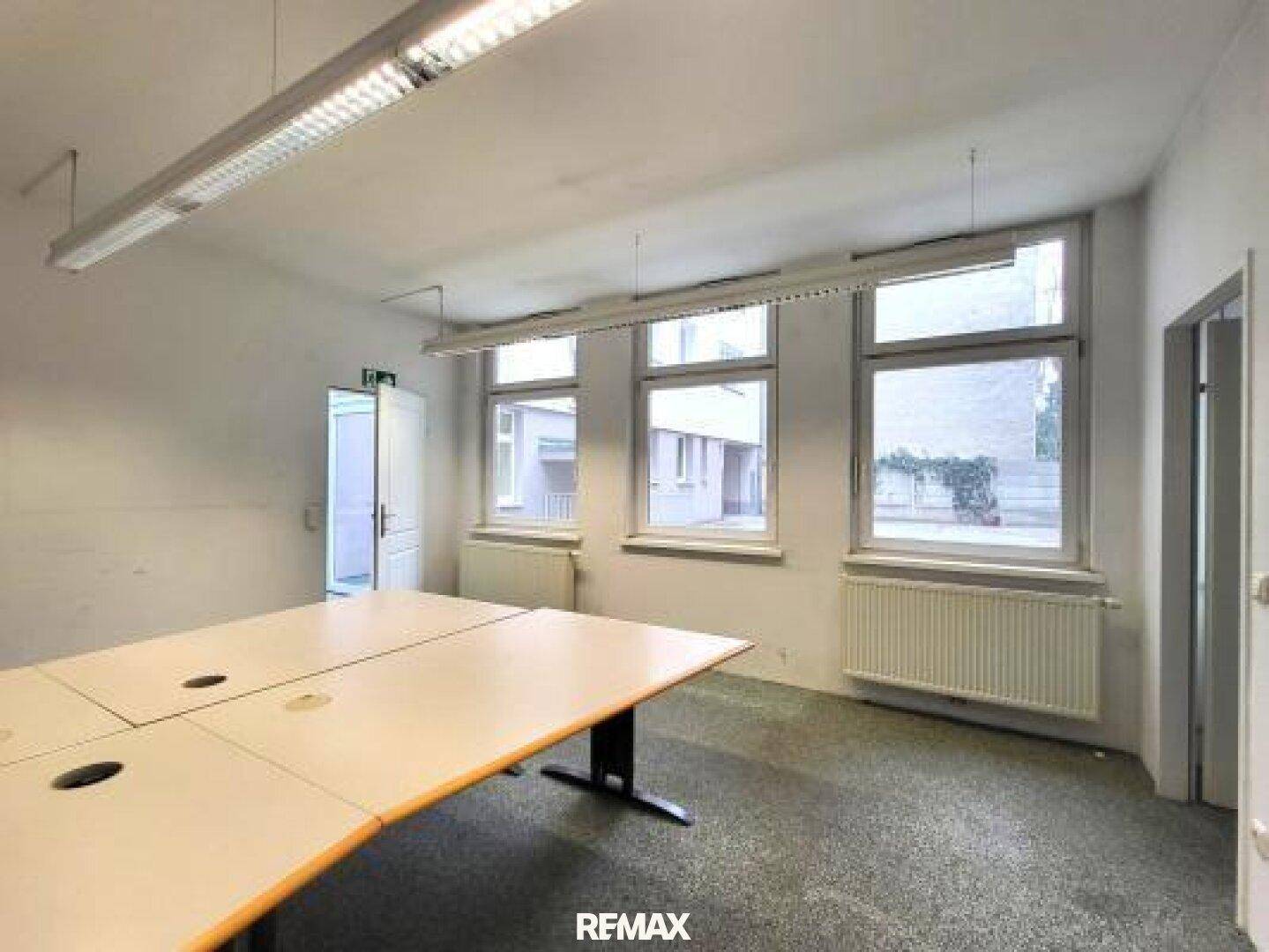 Leerer Büroraum mit mehreren Fenstern, einem großen Tisch und einer Tür.